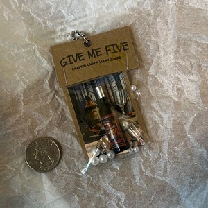 K&ouml;nnte beinhalten: Ein kleiner brauner Papierbeutel mit dem Aufdruck "GIVE ME FIVE CRAFTED UNDER LUCKY STARS" auf der Vorderseite. Der Beutel enth&auml;lt eine Miniaturflasche Alkohol, ein Miniaturglas und wei&szlig;e Perlen.