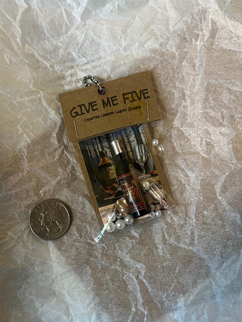K&ouml;nnte beinhalten: Ein kleiner brauner Papierbeutel mit dem Aufdruck "GIVE ME FIVE CRAFTED UNDER LUCKY STARS" auf der Vorderseite. Der Beutel enth&auml;lt eine Miniaturflasche Alkohol, ein Miniaturglas und wei&szlig;e Perlen.