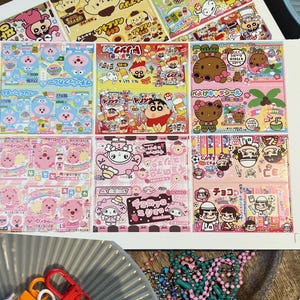 May include: A collection of colorful stickers featuring cartoon characters and Japanese text. The stickers are arranged on a white surface, with a variety of designs and colors. Also visible are keychains and a small bear-shaped pastry.