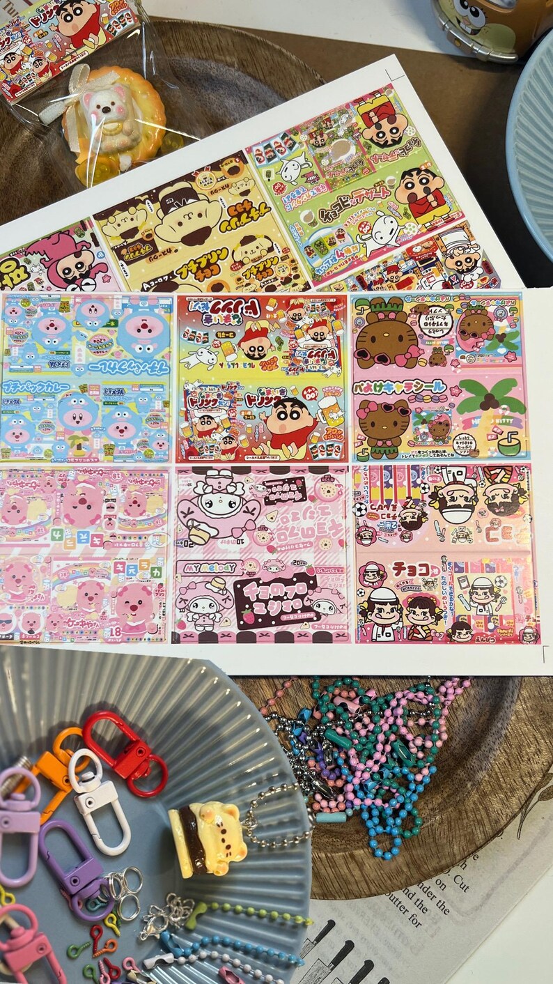 May include: A collection of colorful stickers featuring cartoon characters and Japanese text. The stickers are arranged on a white surface, with a variety of designs and colors. Also visible are keychains and a small bear-shaped pastry.