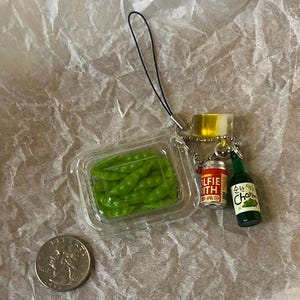 K&ouml;nnte beinhalten: Ein gr&uuml;ner Plastik-Schl&uuml;sselanh&auml;nger mit einem durchsichtigen Plastikbeh&auml;lter mit gr&uuml;nen Bohnen, einer rot-wei&szlig;en Dose mit der Aufschrift "Wolfie with IPA" und einer gr&uuml;nen Glasflasche mit der Aufschrift "Charm" in koreanischer Sprache.