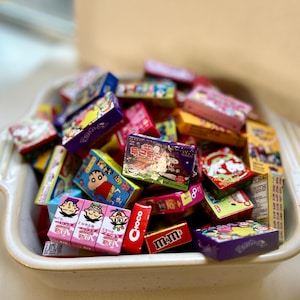 Può includere: Un piatto in ceramica bianca pieno di scatole di caramelle in miniatura colorate. Le scatole presentano vari personaggi dei cartoni animati e loghi, tra cui "Choco", "M&M's" e caratteri giapponesi.