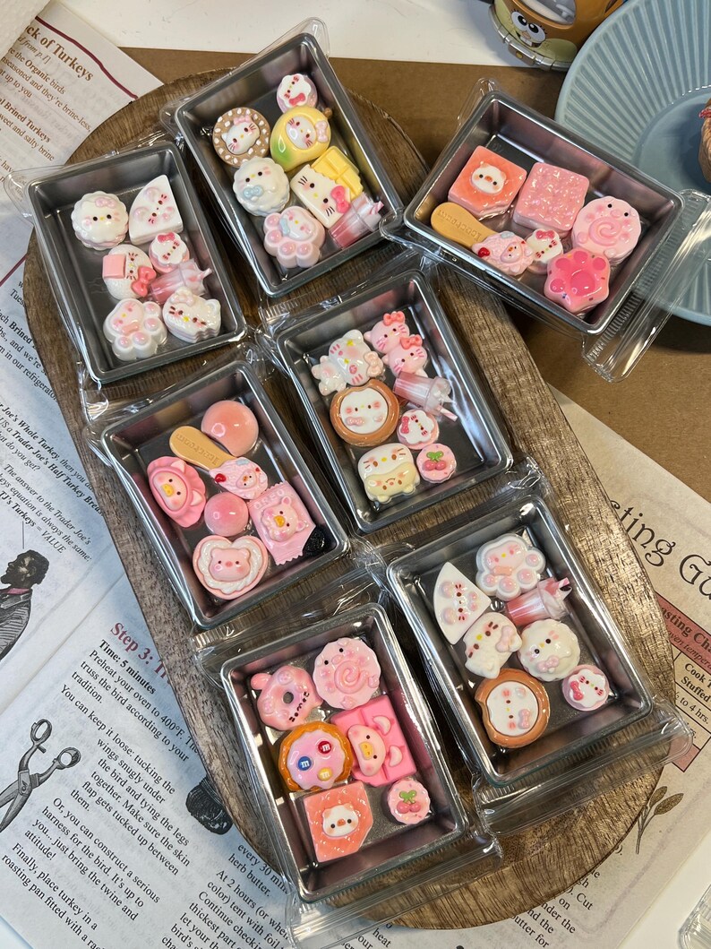 May include: Assorted miniature, colorful, decorative resin pieces in small metal trays. The trays are arranged on a wooden surface. The pieces include various shapes like animals, desserts, and other cute designs. The pieces are packaged in clear plastic.