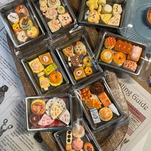 May include: A collection of miniature food items, including pastries, fruits, and other treats, arranged in clear plastic containers. The food items are brightly colored and detailed, with various shapes and designs. The image is taken from above.