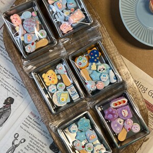 May include: A collection of miniature, colorful, decorative food-themed charms in clear plastic containers. The charms include various shapes like animals, utensils, and food items, in shades of pink, blue, yellow, and purple. They are arranged on a wooden tray.