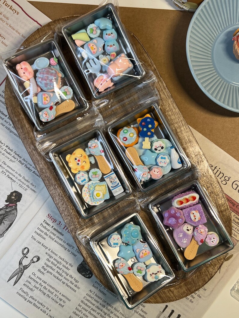 May include: A collection of miniature, colorful, decorative food-themed charms in clear plastic containers. The charms include various shapes like animals, utensils, and food items, in shades of pink, blue, yellow, and purple. They are arranged on a wooden tray.