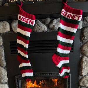 Peut inclure: Deux chaussettes de Noël rayées rouge, blanc et vert avec les noms "Lauren" et "Bennett" accrochées au-dessus d'une cheminée avec un feu qui brûle à l'intérieur.