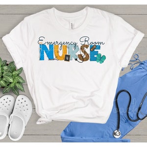 May include: White t-shirt with the text "Emergency Room Nurse" in blue, yellow, and brown letters. The letters are decorated with medical symbols and tools. The shirt is displayed on a wooden surface with a pair of white clogs, a blue scrub top, and a stethoscope.