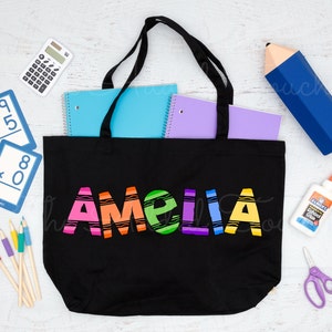 May include: Black canvas tote bag with the name "Amelia" written in rainbow colored crayon-like letters. The bag is filled with school supplies, including notebooks, pencils, and a calculator.