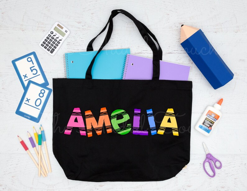 May include: Black canvas tote bag with the name "Amelia" written in rainbow colored crayon-like letters. The bag is filled with school supplies, including notebooks, pencils, and a calculator.