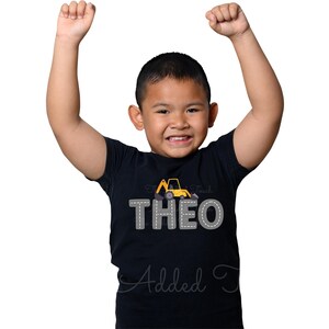 May include: A black t-shirt featuring a yellow construction vehicle graphic and the name "THEO" in grey outlined letters.
