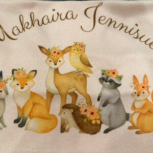 May include: A light pink towel featuring a watercolor illustration of woodland animals, including a deer, fox, owl, raccoon, squirrel, hedgehog, and rabbit, each adorned with floral crowns. The text "Makhaira Jennisiue" is arched across the top.