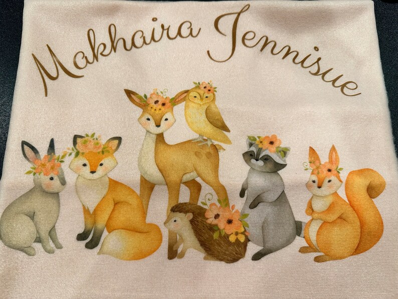 May include: A light pink towel featuring a watercolor illustration of woodland animals, including a deer, fox, owl, raccoon, squirrel, hedgehog, and rabbit, each adorned with floral crowns. The text "Makhaira Jennisiue" is arched across the top.