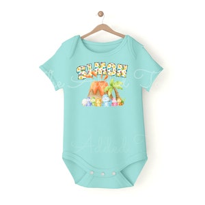 K&ouml;nnte beinhalten: Ein hellblauer Baby-Strampler mit einem Vulkan und Dinosauriern. Der Text "SIMON" ist auf dem Strampler aufgedruckt.