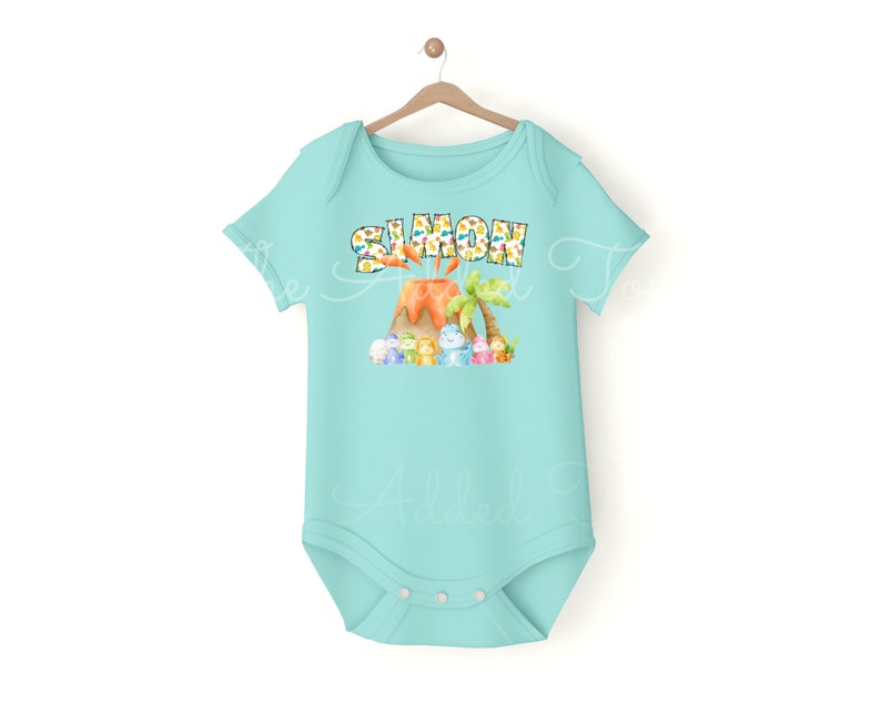 K&ouml;nnte beinhalten: Ein hellblauer Baby-Strampler mit einem Vulkan und Dinosauriern. Der Text "SIMON" ist auf dem Strampler aufgedruckt.