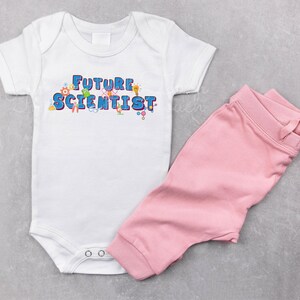 Pu&ograve; includere: Un body bianco per neonato con il testo "FUTURE SCIENTIST" in lettere blu e grafiche colorate a tema scientifico. Il body &egrave; abbinato a pantaloni rosa.