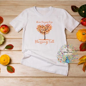 May include: White t-shirt with the text "When Prayers Rise Blessings Fall" and a tree design with autumn leaves. The shirt is surrounded by pumpkins, gourds, apples, and autumn leaves on a wooden surface.