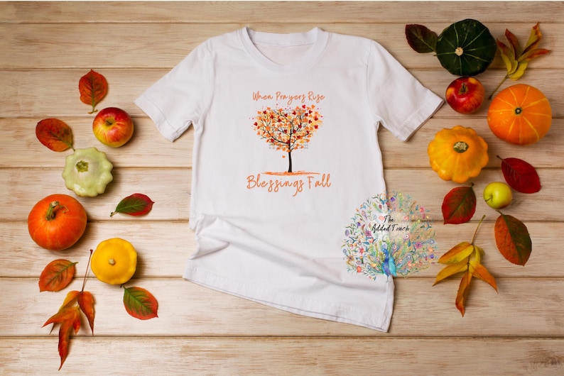 May include: White t-shirt with the text "When Prayers Rise Blessings Fall" and a tree design with autumn leaves. The shirt is surrounded by pumpkins, gourds, apples, and autumn leaves on a wooden surface.