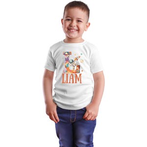May include: White t-shirt with a colorful letter L design featuring an astronaut, planets, and a rocket. The name "LIAM" is printed in orange below the letter.