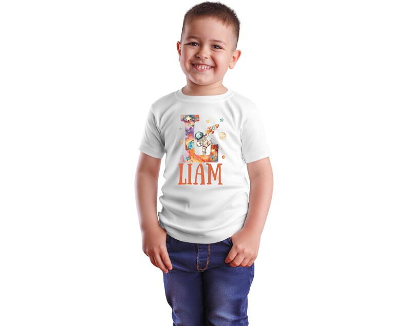 May include: White t-shirt with a colorful letter L design featuring an astronaut, planets, and a rocket. The name "LIAM" is printed in orange below the letter.