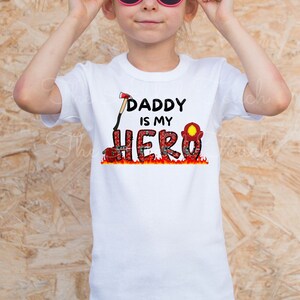 Op de afbeelding: Een wit T-shirt met een rode en zwarte afbeelding waarop staat "Daddy is my Hero". De afbeelding toont een brandweerhelm, een bijl en vlammen.