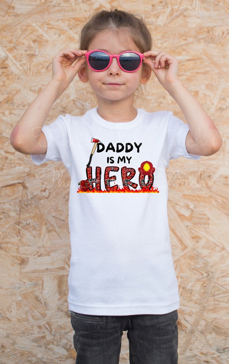 Op de afbeelding: Een wit T-shirt met een rode en zwarte afbeelding waarop staat "Daddy is my Hero". De afbeelding toont een brandweerhelm, een bijl en vlammen.