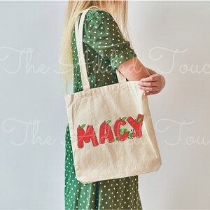 May include: A white canvas tote bag with the name "MACY" written in red letters with green leaves and a red caterpillar on the bag.