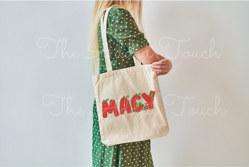 May include: A white canvas tote bag with the name "MACY" written in red letters with green leaves and a red caterpillar on the bag.