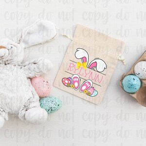 May include: A white bunny-shaped Easter gift bag with a pink bunny design and the name "RYLYNN" printed on it. The bag is filled with colorful Easter eggs.