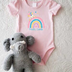 May include: Pink baby onesie with a rainbow and a unicorn peeking out. The onesie has the names "Felicity Camille" printed below the rainbow.