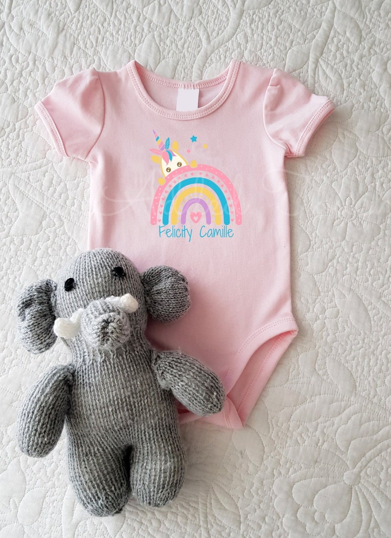 May include: Pink baby onesie with a rainbow and a unicorn peeking out. The onesie has the names "Felicity Camille" printed below the rainbow.