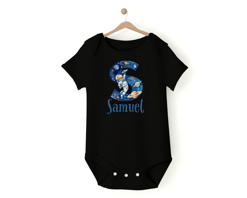 May include: Black baby onesie with a blue and white astronaut graphic in the shape of the letter S. The onesie has the name "Samuel" printed below the graphic.