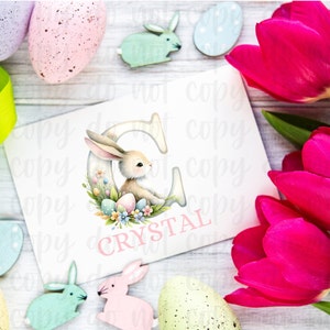 May include: A white card with a pink bunny rabbit inside a letter C. The letter C is surrounded by pastel colored Easter eggs and flowers. The card says "CRYSTAL" in pink letters.