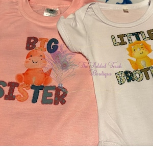 May include: Two infant shirts, one pink and one white, with a dinosaur and a lion graphic. The pink shirt says "Big Sister" and the white shirt says "Little Brother".