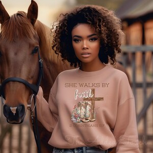May include: A woman wearing a light brown sweatshirt with the text "She Walks By Faith" and a cross with cowboy boots underneath. The text "2 Corinthians 5:7" is below the boots.