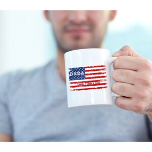 May include: White ceramic mug with a red, white, and blue American flag design. The text "DADA" is printed above the flag, and the names "Charlie, Claire, & Syra" are printed below the flag.
