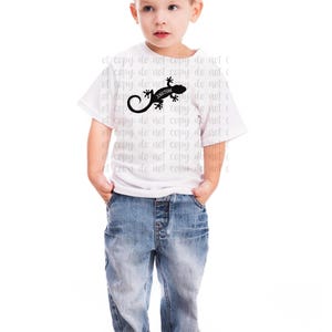 Pu&ograve; includere: Un bambino indossa una maglietta bianca con una grafica di lucertola nera e il nome "Jonathan". Il bambino indossa anche jeans blu. L'immagine &egrave; su sfondo bianco.