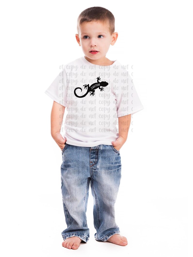 Pu&ograve; includere: Un bambino indossa una maglietta bianca con una grafica di lucertola nera e il nome "Jonathan". Il bambino indossa anche jeans blu. L'immagine &egrave; su sfondo bianco.