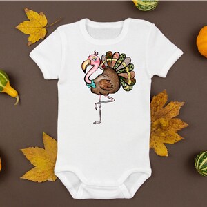 May include: White baby bodysuit featuring a whimsical illustration of a flamingo-turkey hybrid. The design includes a pink head, brown body, and colorful tail feathers. The bodysuit is surrounded by pumpkins and fall leaves.