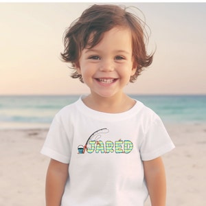 May include: A white t-shirt featuring a colourful graphic of a fishing rod, bucket, and the name "JARED". The shirt is being worn by a young child standing on a beach.