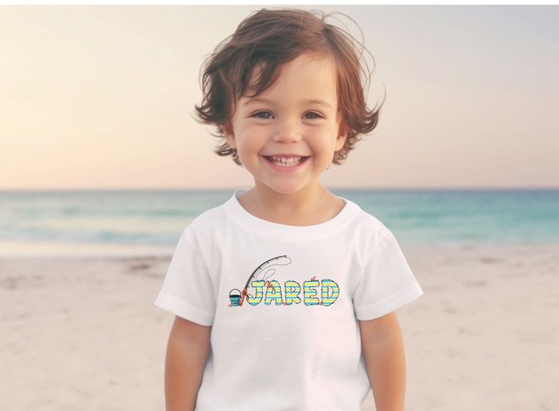 May include: A white t-shirt featuring a colourful graphic of a fishing rod, bucket, and the name "JARED". The shirt is being worn by a young child standing on a beach.