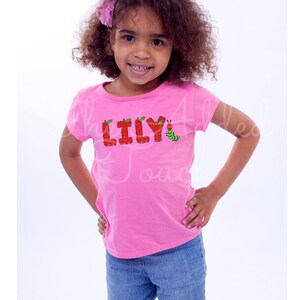 May include: A young girl wearing a pink t-shirt with the name "LILY" spelled out in red letters with green leaves and an apple on each letter. A green caterpillar with a red head is to the right of the name.