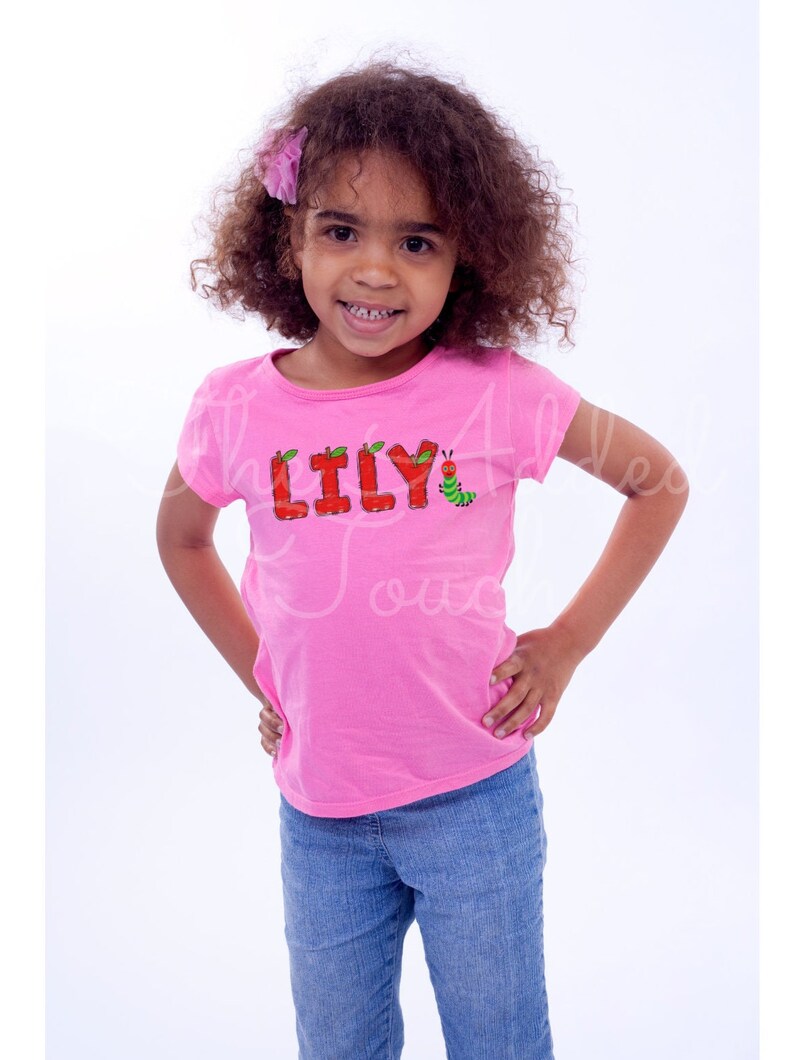May include: A young girl wearing a pink t-shirt with the name "LILY" spelled out in red letters with green leaves and an apple on each letter. A green caterpillar with a red head is to the right of the name.