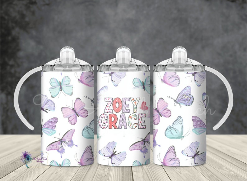May include: Three white stainless steel cups with clear lids and handles. The cups have a butterfly pattern with pink, purple, and blue butterflies. The middle cup has the names "ZOEY & GRACE" in pink, red, and white letters.