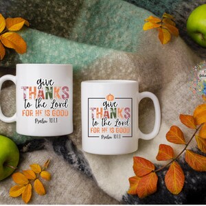 May include: Two white ceramic mugs with a fall-themed design. The mugs feature the text "give thanks to the Lord for he is good" with a pumpkin and fall leaves. The mugs are on a brown and white blanket with fall leaves.
