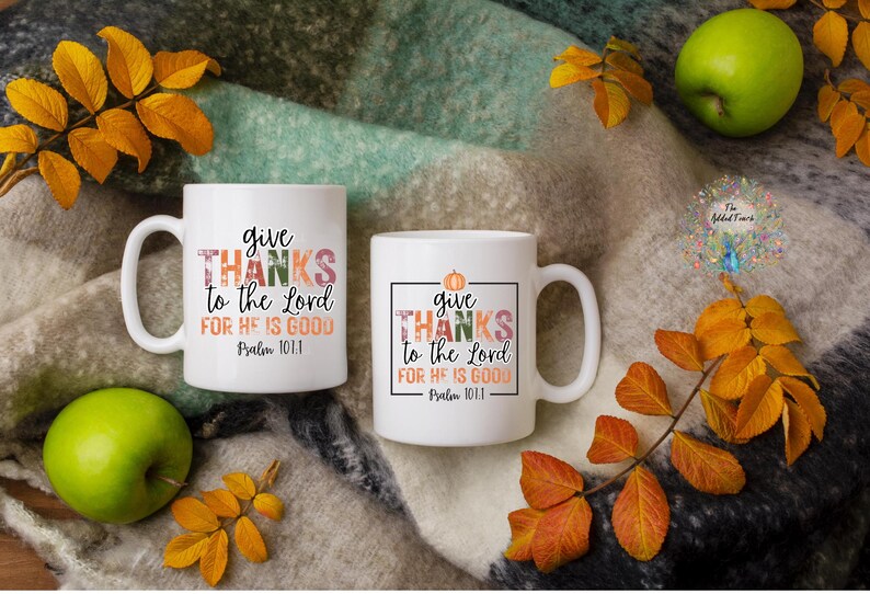 May include: Two white ceramic mugs with a fall-themed design. The mugs feature the text "give thanks to the Lord for he is good" with a pumpkin and fall leaves. The mugs are on a brown and white blanket with fall leaves.