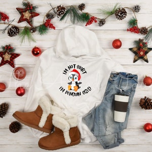 May include: A white hooded sweatshirt with a penguin graphic wearing a Santa hat and the text "I'm not short, I'm penguin sized." The image also includes blue jeans, brown boots, and holiday decorations.