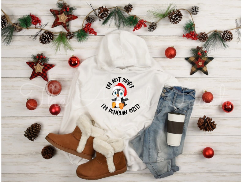 May include: A white hooded sweatshirt with a penguin graphic wearing a Santa hat and the text "I'm not short, I'm penguin sized." The image also includes blue jeans, brown boots, and holiday decorations.