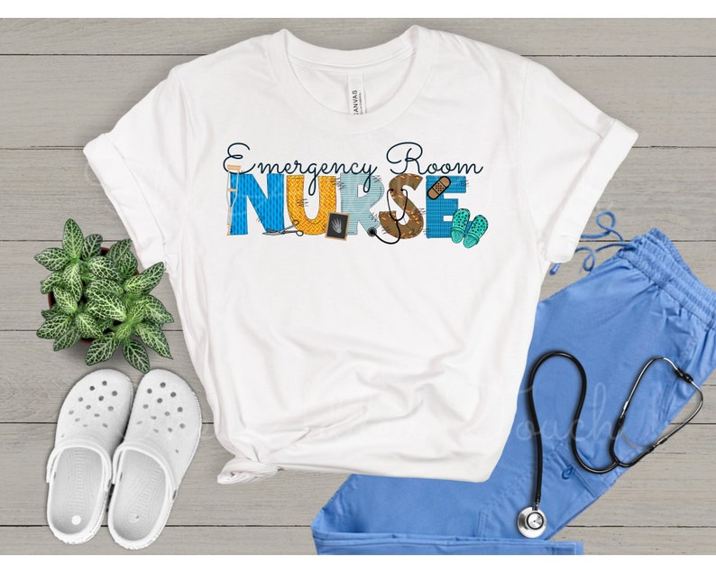 May include: White t-shirt with the text "Emergency Room Nurse" in blue, yellow, and brown letters. The letters are decorated with medical symbols and tools. The shirt is displayed on a wooden surface with a pair of white clogs, a blue scrub top, and a stethoscope.