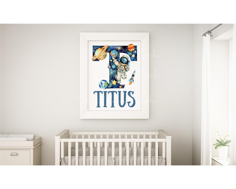 May include: A white framed print with a blue and black letter T filled with a space scene. An astronaut is holding a planet and a rocket is flying by. The name Titus is written in blue below the letter.
